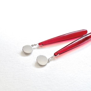 Op de afbeelding: Een paar rode, druppelvormige oorbellen met zilverkleurige, cirkelvormige accenten. De oorbellen worden gepresenteerd op een wit oppervlak, waarbij hun levendige kleur en strakke ontwerp worden benadrukt. Een stijlvol accessoire.