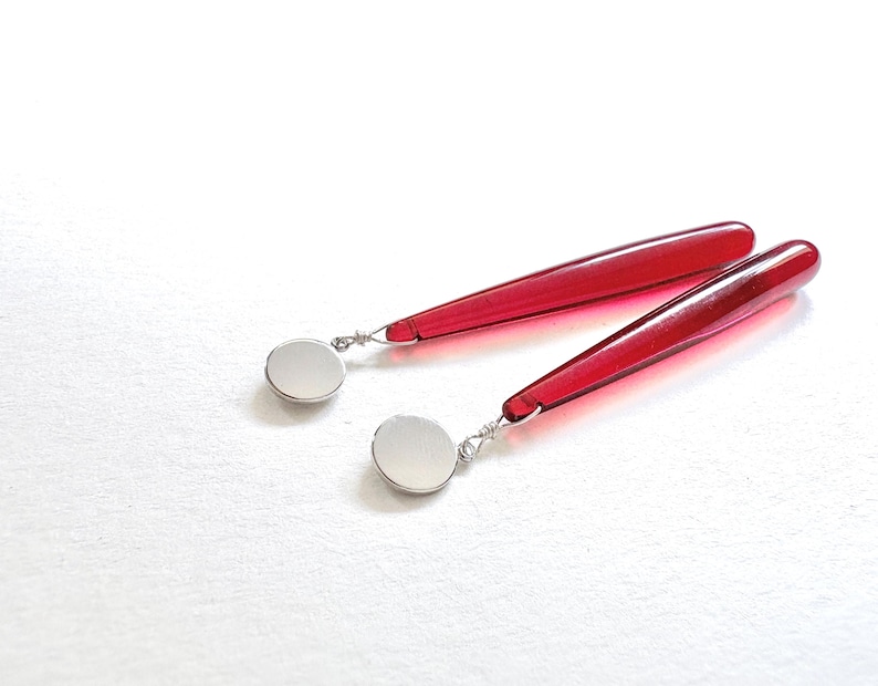 Op de afbeelding: Een paar rode, druppelvormige oorbellen met zilverkleurige, cirkelvormige accenten. De oorbellen worden gepresenteerd op een wit oppervlak, waarbij hun levendige kleur en strakke ontwerp worden benadrukt. Een stijlvol accessoire.