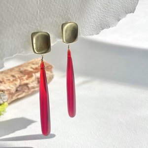 Op de afbeelding: Een paar goudkleurige vierkante oorstekers met lange, druppelvormige, doorschijnende rode hangers. De oorbellen zijn opgehangen aan dunne draadjes en worden gepresenteerd tegen een witte achtergrond met natuurlijk licht en schaduwen.