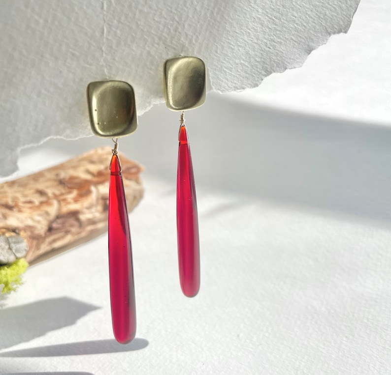 Op de afbeelding: Een paar goudkleurige vierkante oorstekers met lange, druppelvormige, doorschijnende rode hangers. De oorbellen zijn opgehangen aan dunne draadjes en worden gepresenteerd tegen een witte achtergrond met natuurlijk licht en schaduwen.