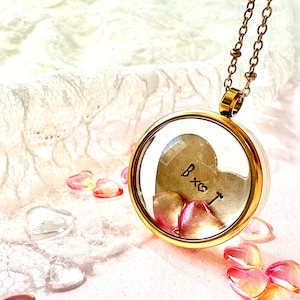 Può includere: Una collana con un medaglione dorato e un fronte in vetro trasparente, che rivela un ciondolo a forma di cuore con le lettere "B x ♡". Il medaglione è riempito di piccole perline a forma di cuore. Anche la catena della collana è dorata.