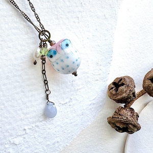 Op de afbeelding: Een handgemaakte uil hanger ketting. De uil bedel is wit met blauwe stippen, roze accenten en blauwe ogen. De ketting heeft een bronzen ketting, een blauwe druppel bedel en kleine parel en groene kraal accenten.