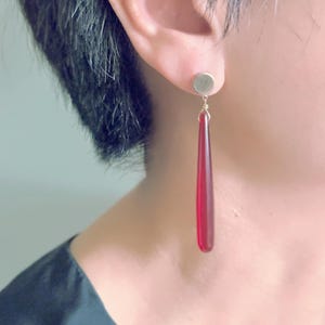 Op de afbeelding: Een enkele oorbel met een kleine, ronde, goudkleurige stud. Een lange, druppelvormige, doorschijnende rode hanger hangt aan de stud. De oorbel wordt op het oor gedragen. De achtergrond is wazig.