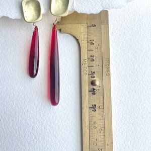 Op de afbeelding: Oorbellen met goudkleurige vierkante bovenstukken en langwerpige, druppelvormige rode hangers. De oorbellen worden gemeten met een messing liniaal, met een lengte van ongeveer 6,35 cm. Gepresenteerd op een witte achtergrond.