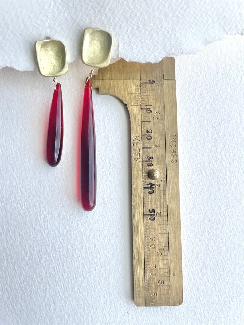 Op de afbeelding: Oorbellen met goudkleurige vierkante bovenstukken en langwerpige, druppelvormige rode hangers. De oorbellen worden gemeten met een messing liniaal, met een lengte van ongeveer 6,35 cm. Gepresenteerd op een witte achtergrond.