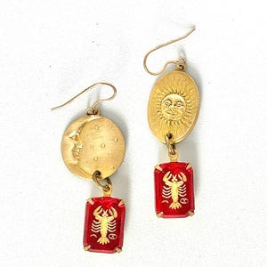 Könnte beinhalten: Goldfarbene Ohrringe mit einem Sonnen- und Mondmotiv und einem roten Glasanhänger mit einem goldenen Krebs-Sternzeichen.