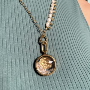 May include: A gold chain necklace with a pearl and spike pendant. The pendant features a round, clear glass orb with a gold sun face design and small, clear crystals inside.