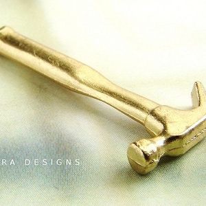 Puede incluir: Un collar con un colgante de martillo de color dorado. El martillo está detallado con una superficie texturizada y una garra curva.
