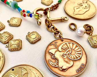 May include: A gold necklace with a charm of the zodiac sign Aries, with the word "Aries" inscribed on the charm. The necklace also features a variety of other charms, including letters, a pearl, and a small anchor.