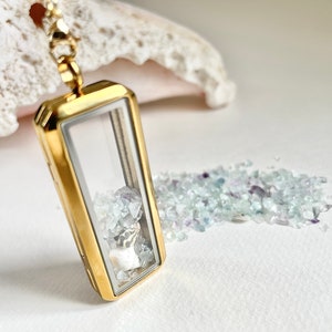 Puede incluir: Un collar de oro con un dije de cristal transparente lleno de pequeños cristales azules y morados. El dije está abierto y los cristales son visibles.