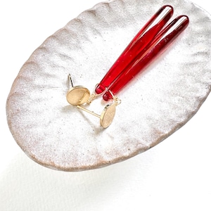 Op de afbeelding: Goudkleurige oorstekers met een klein, rond ontwerp en twee lange, rode glazen staven. De oorbellen en staven worden gepresenteerd op een getextureerde, ovaalvormige, gebroken witte keramische schaal.