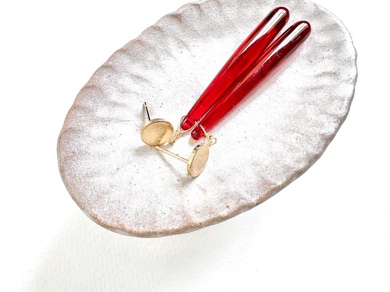 Op de afbeelding: Goudkleurige oorstekers met een klein, rond ontwerp en twee lange, rode glazen staven. De oorbellen en staven worden gepresenteerd op een getextureerde, ovaalvormige, gebroken witte keramische schaal.