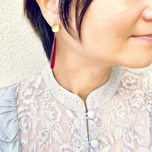 Op de afbeelding: Close-up van een persoon die goudkleurige vierkante oorstekers draagt met lange, druppelvormige rode hangers. De persoon draagt een lichtblauwe kanten blouse.