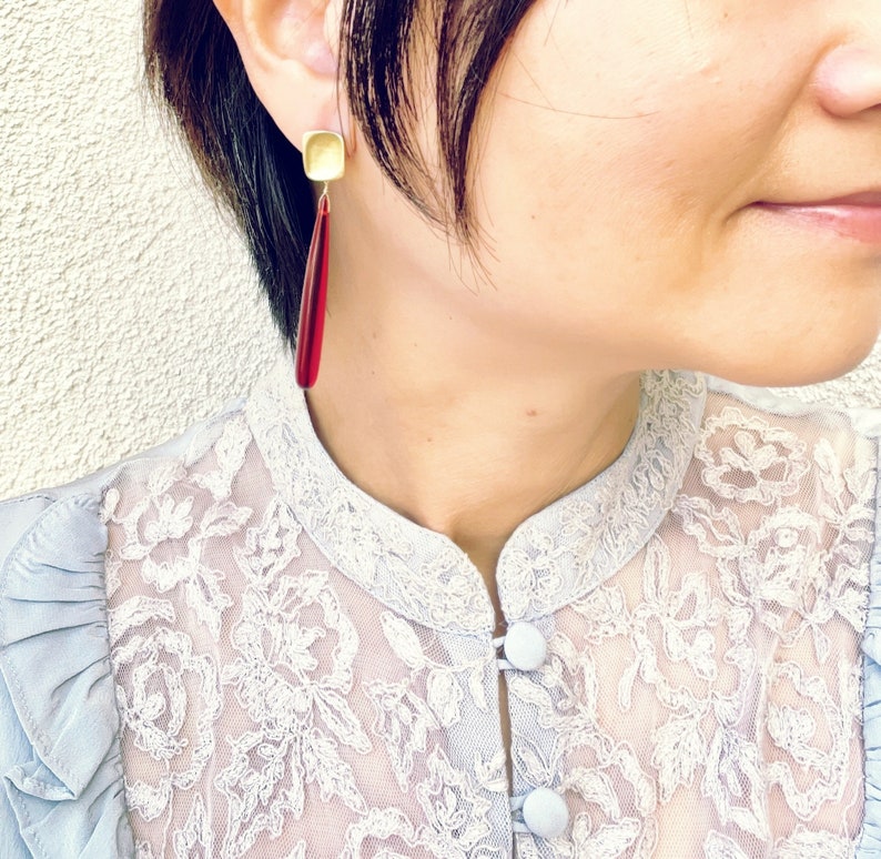 Op de afbeelding: Close-up van een persoon die goudkleurige vierkante oorstekers draagt met lange, druppelvormige rode hangers. De persoon draagt een lichtblauwe kanten blouse.