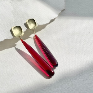 Op de afbeelding: Oorbellen met vierkante, goudkleurige studs en lange, druppelvormige hangers in een dieprode tint. De oorbellen worden gepresenteerd op een witte ondergrond, met schaduwen die diepte toevoegen. De sieraden hebben een modern, minimalistisch ontwerp.