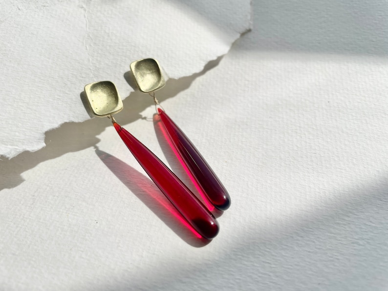 Op de afbeelding: Oorbellen met vierkante, goudkleurige studs en lange, druppelvormige hangers in een dieprode tint. De oorbellen worden gepresenteerd op een witte ondergrond, met schaduwen die diepte toevoegen. De sieraden hebben een modern, minimalistisch ontwerp.