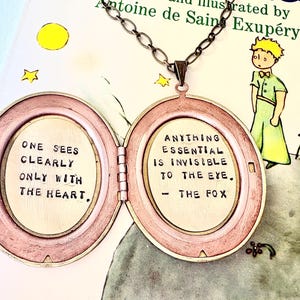 Può includere: Una collana con un medaglione aperto in stile vintage, con una catena in bronzo. Il medaglione ovale ha una cornice rosa-bronzo e contiene due citazioni: "SI VEDE BENE SOLO CON IL CUORE" e "L'ESSENZIALE È INVISIBILE AGLI OCCHI. - LA VOLPE."