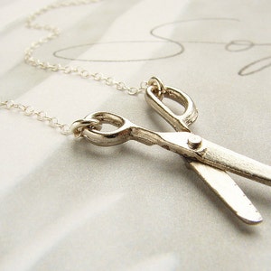 May include: A silver necklace with a silver scissor pendant. The necklace is on a white surface.
