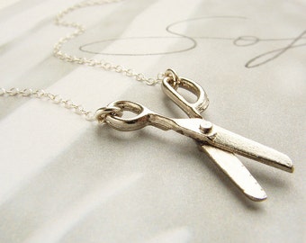 Op de afbeelding: Een zilveren ketting met een zilveren schaar hanger. De ketting ligt op een wit oppervlak.