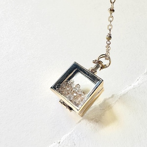 Peut inclure: Un collier pendentif carré doré avec une face en verre transparent, rempli de petits cristaux étincelants. Le pendentif est attaché à une chaîne délicate. Le collier est présenté sur un fond blanc.