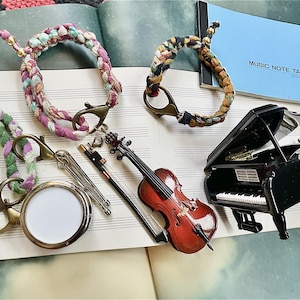 Puede incluir: Primer plano de un conjunto de instrumentos musicales en miniatura, que incluyen un violonchelo, un piano y una batería. Los instrumentos están hechos de metal y madera y están pintados en colores realistas. Los instrumentos están dispuestos sobre una superficie blanca con una libreta de notas musicales en el fondo.