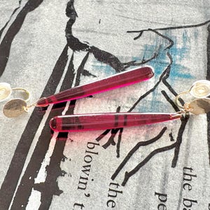 Op de afbeelding: Een paar oorhangers met een goudkleurige oorbel met een kleine, getextureerde schijf en een lange, transparante, magenta-kleurige, rechthoekige hanger. De oorbellen liggen op een patroon oppervlak.