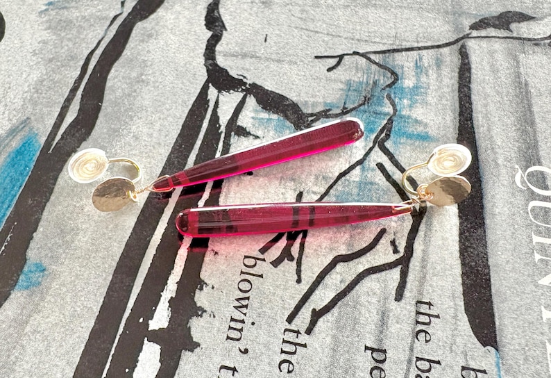 Op de afbeelding: Een paar oorhangers met een goudkleurige oorbel met een kleine, getextureerde schijf en een lange, transparante, magenta-kleurige, rechthoekige hanger. De oorbellen liggen op een patroon oppervlak.