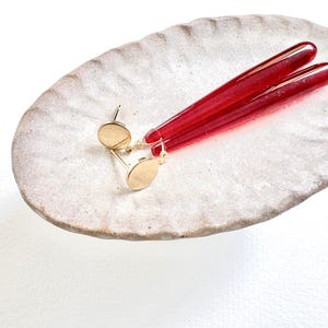 Op de afbeelding: Goudkleurige oorstekers met lange, rode, cilindervormige hangers. De oorbellen worden gepresenteerd op een getextureerde, off-white, ovale keramische schaal. De rode hangers hebben een levendige, doorschijnende kleur.