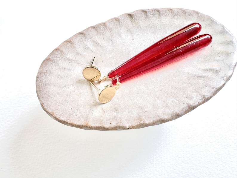 Op de afbeelding: Goudkleurige oorstekers met lange, rode, cilindervormige hangers. De oorbellen worden gepresenteerd op een getextureerde, off-white, ovale keramische schaal. De rode hangers hebben een levendige, doorschijnende kleur.