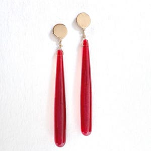 Op de afbeelding: Een paar oorhangers met goudkleurige cirkelvormige studs en lange, druppelvormige, dieprode glazen hangers. De oorbellen hangen verticaal tegen een witte achtergrond. Het ontwerp is eenvoudig en elegant.