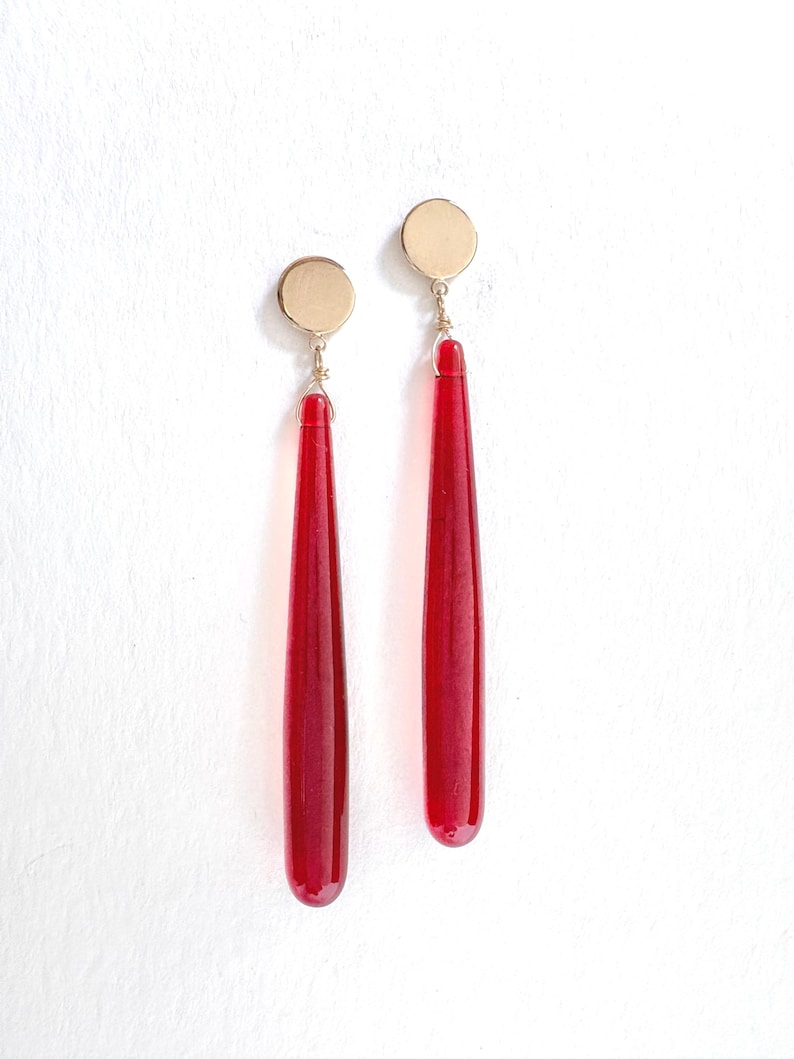 Op de afbeelding: Een paar oorhangers met goudkleurige cirkelvormige studs en lange, druppelvormige, dieprode glazen hangers. De oorbellen hangen verticaal tegen een witte achtergrond. Het ontwerp is eenvoudig en elegant.