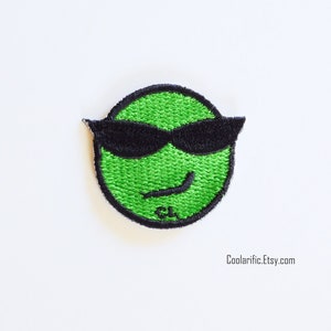 Puede incluir: Parche bordado verde con una cara sonriente que lleva gafas de sol negras. La cara tiene una leve sonrisa y las gafas de sol están inclinadas.