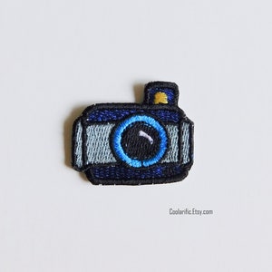 May include: Blue and gray embroidered iron-on patch of a camera with a yellow flash.