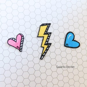 May include: Three embroidered iron-on patches: a pink heart, a yellow lightning bolt, and a blue heart. The patches are on a white hexagonal background.