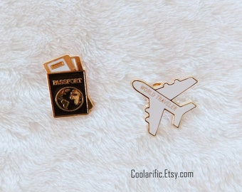 Könnte beinhalten: Zwei goldene Emaille-Anstecknadeln. Eine Nadel zeigt einen Reisepass mit einem Globus und dem Wort "PASSPORT" darauf. Die andere Nadel zeigt ein weißes Flugzeug mit den Worten "WORLD TRAVELER" darauf.