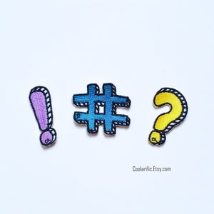 May include: Three embroidered iron-on patches in the shape of an exclamation mark, hashtag, and question mark. The exclamation point is purple, the hashtag is blue, and the question mark is yellow.