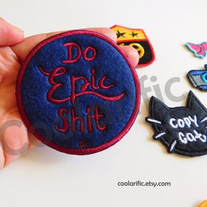 以下が含まれることがあります： マロン色の縁取りのある、紺色の丸い刺繍パッチ。パッチには「Do Epic Shit」という白い文字が書かれています。