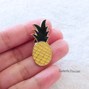 May include: A gold-coloured pineapple-shaped enamel pin with a yellow body and black top. The pin is held in a hand against a white background. The Etsy shop URL is visible at the bottom.