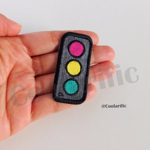 Puede incluir: Un parche bordado negro y gris de un semáforo con luces rojas, amarillas y verdes.