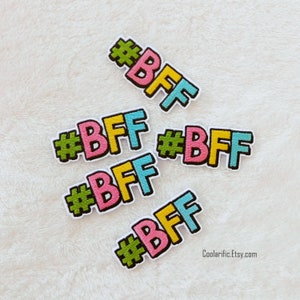Puede incluir: Cinco parches bordados para planchar con el texto "#BFF" en diferentes colores. Los parches están sobre una superficie blanca y peluda.