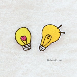 May include: Two yellow embroidered iron-on patches shaped like light bulbs. One light bulb has a pink heart inside. The text "Coolariific.Etsy.com" is in the bottom right corner of the image.