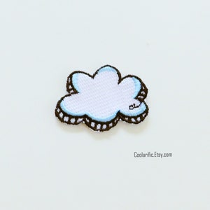 Peut inclure: Un nuage blanc avec un contour bleu et une bordure noire à pois. Le nuage est brodé sur un tissu blanc.