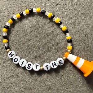 May include: A beaded bracelet with the words "HOIST THE" spelled out in white letter beads. The bracelet features a mix of black, yellow, and clear beads, and is accented with a small orange and white traffic cone charm.