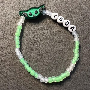 May include: A green and white beaded bracelet with a green Yoda charm and the word "YODA" spelled out in white beads.