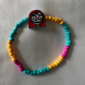 Puede incluir: Un brazalete de cuentas de colores con un dije de personaje rojo, blanco y azul. El dije tiene una cara de dibujos animados con una expresión traviesa. El brazalete está hecho de pequeñas cuentas redondas en tonos de amarillo, rosa y turquesa.