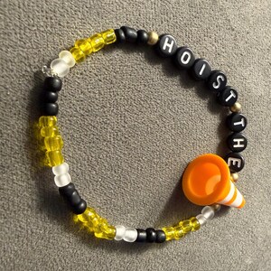 May include: A beaded bracelet with the words "HOIST THE" spelled out in black letter beads. The bracelet features yellow, white, and black beads, and an orange traffic cone charm.
