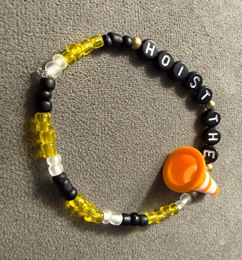May include: A beaded bracelet with the words "HOIST THE" spelled out in black letter beads. The bracelet features yellow, white, and black beads, and an orange traffic cone charm.