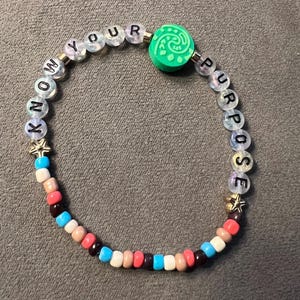 May include: A beaded bracelet with the words "KNOW YOUR PURPOSE" spelled out in black letters on white beads. The bracelet also features a green bead with a swirl design and colorful beads in shades of red, orange, brown, blue, and white.