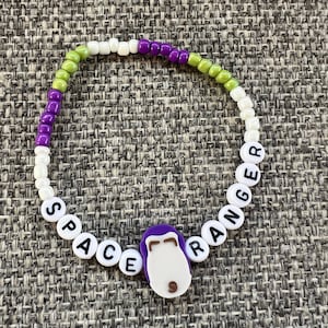 May include: White beaded bracelet with the words "SPACE RANGER" spelled out in black letters. The bracelet has green and purple beads and a purple and white cartoon character charm.