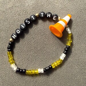 May include: A beaded bracelet with the words "HOIST THE" spelled out in black letter beads. The bracelet features a small orange and white traffic cone charm, and is strung with yellow, black, and white beads.